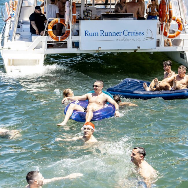 People are swimming and floating on inflatables in the water near a boat. The boat has a banner that reads "Rum Runner Cruises," and more people are onboard. Some are standing, and others are seated, enjoying drinks from the onboard tiki bar. The lively water scene brims with various activities.