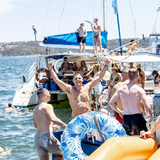 A group of people are enjoying a sunny day on a boat. Some lounge on the deck, while others swim in the water. One person stands with their arms raised in excitement, holding a blue inflatable pool ring. A large inflatable slide extends into the sparkling water, adding to the fun.