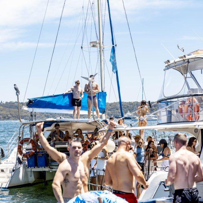 A group of people are enjoying a sunny day on and around sailboats and motorboats anchored close together. Some are on the decks, while others are in the water or standing on the boats. A man in swim trunks raises his arms in excitement, celebrating the perfect boating adventure.