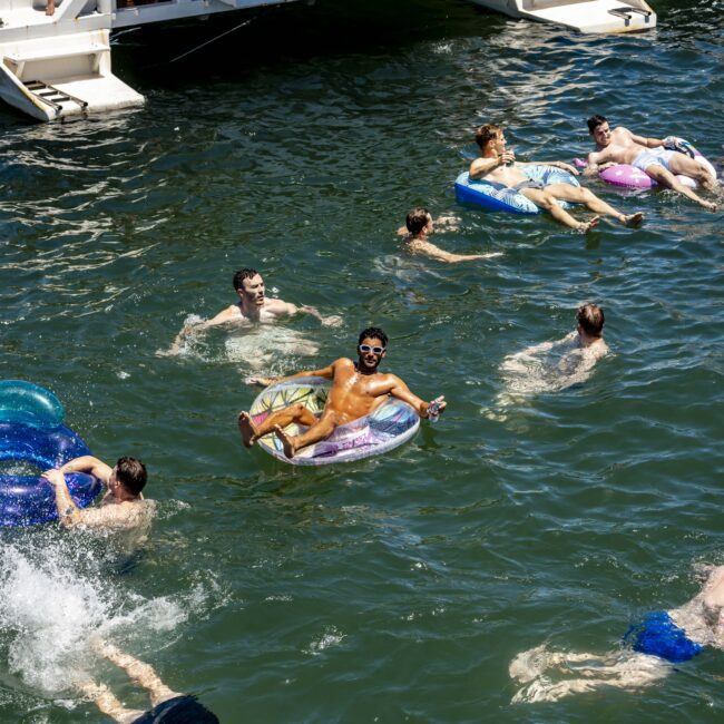 People enjoy a sunny day in the water, swimming and relaxing on inflatables near a boat labeled "Rum Runner Cruises." The scene is lively, with individuals engaging in different activities, including diving into the water. The Yacht Social Club logo is visible on some of the inflatables.