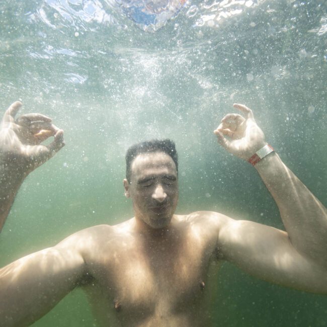 A man is underwater with both arms raised and fingers forming the "OK" sign. He is calm, with closed eyes and a slight smile, indicating a sense of serenity. Light from above creates mesmerizing patterns in the water, enhancing the tranquil scene. A logo in the bottom right corner says “The Yacht Social Club.”
