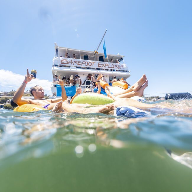 People float on inflatable rings in the water near a large boat named "Barefoot Explorer." The boat, with people socializing on its decks, is anchored against a clear blue sky. The image has a watermark in the bottom right corner that reads "Yacht Social Club.
