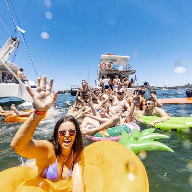 A group of people are enjoying a sunny day on the water, floating on colorful inflatables near several yachts. One person in the foreground waves at the camera, smiling. The vibrant and festive setting is enhanced by a nearby beach, with everyone engaging in the fun.