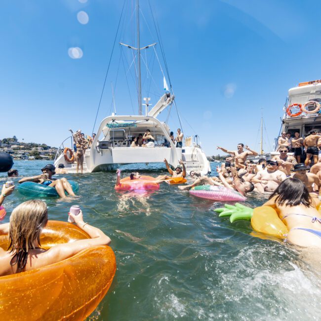 A lively gathering on the water, with people on inflatable floaties near two anchored boats. Participants, in swimsuits, enjoy sunny weather, some in conversation, others splashing in the water. The clear blue sky and distant hills enhance this vibrant and festive scene of summer fun.