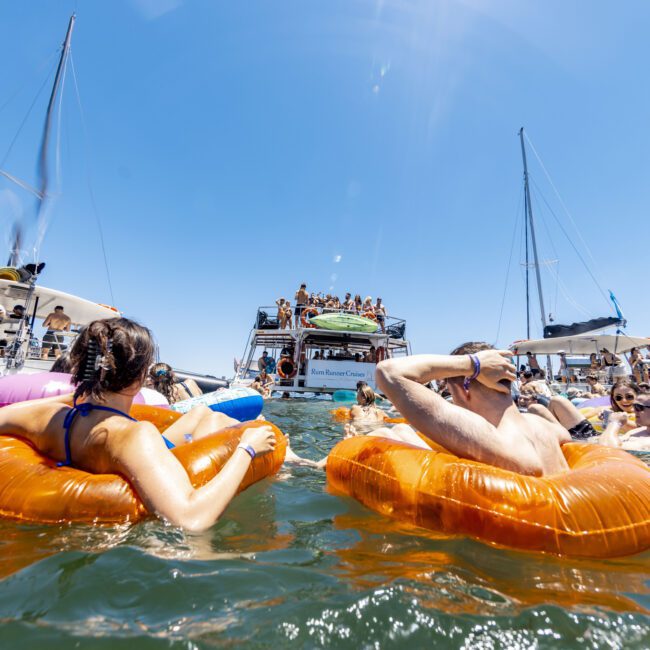 A lively scene of families enjoying a sunny day at sea. Several individuals are floating on inflatable tubes in the water near large yachts. The atmosphere is festive, with more people visible on the boats, some dancing and socializing. The sky is clear and bright.
