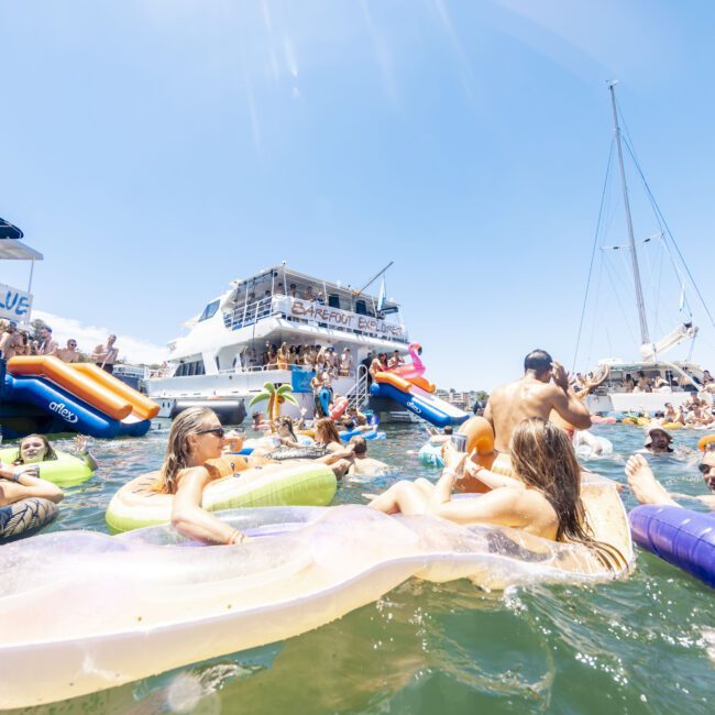 People enjoying a sunny day at a floating party, surrounded by multiple boats and yachts. Many are lounging on colorful inflatables in the water, with music and festivities creating a vibrant atmosphere. The water is crystal clear, and the sky is a bright blue canvas above.