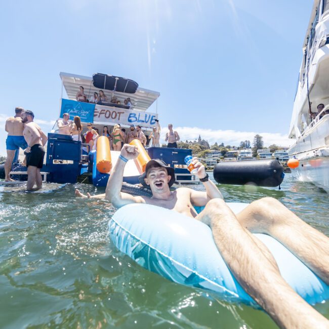 A man lounges on a blue inflatable raft in the water, smiling and holding a drink, surrounded by partygoers on nearby boats. There is a floating bar with a sign reading "Foot Blue" in the background. The atmosphere is lively and festive under a clear blue sky, making it feel like an adventure.