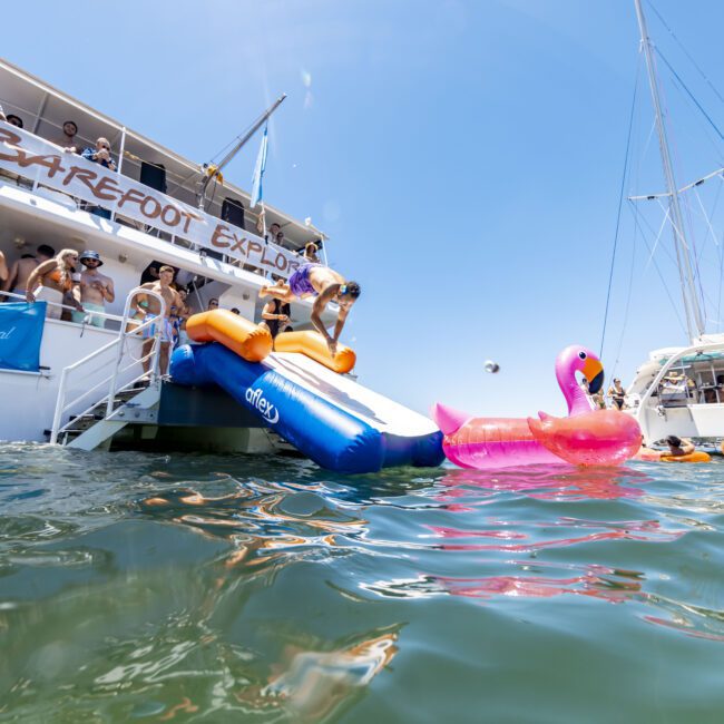 A large boat named "Carefree Explorer" is anchored in the water with people enjoying a sunny day. Some are sliding down an inflatable slide into the water, where there's a pink flamingo float. Additional boats and more people on jet skis can be seen in the background.