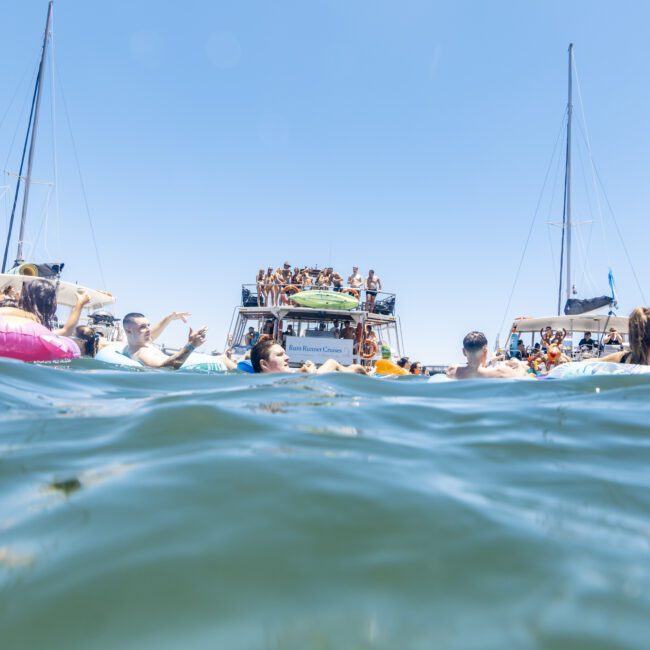 People are enjoying a sunny day on the water, swimming near several anchored boats. The boats are filled with partygoers, and one central boat has a crowded upper deck, contributing to the festive scene. The bright blue skies and greenish water enhance the lively atmosphere. The logo "Yacht Social Club" is visible in the bottom right corner.