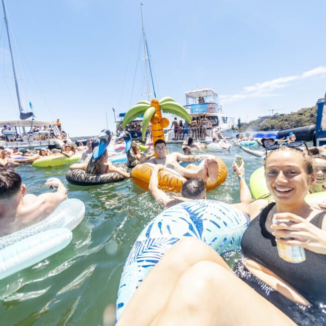 A vibrant scene of people enjoying a sunny day on the water with various inflatable floats. Boats are anchored nearby, and a woman in the foreground is smiling and holding a drink. The background features a clear blue sky as people socialize on brightly colored floats and boats.