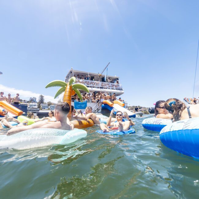 A lively scene shows people relaxing and socializing on inflatable rafts in the water near several yachts. One of the floats features an inflatable palm tree. The sky is clear and sunny, with onlookers visible on the yachts in the background. "Yacht Social Club" is printed in the corner.