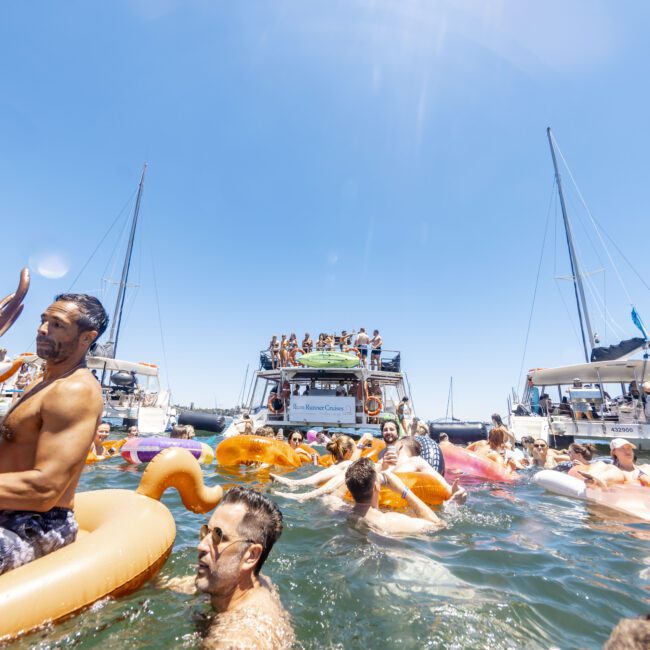 A vibrant outdoor party scene on the water, with numerous people swimming and lounging on various inflatable floats. In the background, three boats are anchored close together, filled with more revelers enjoying the sunny day. The atmosphere is lively and energetic, enhanced by a backdrop of sparkling water.