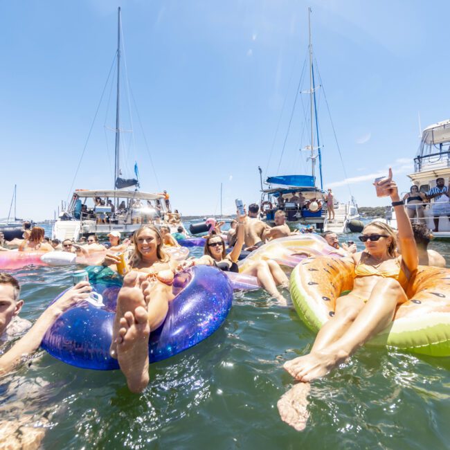 A lively scene of people lounging on colorful inflatable rafts and tubes in the water, surrounded by yachts and boats on a sunny day. The atmosphere is festive, with individuals relaxing, smiling, and enjoying the vibrant ambiance of a beach party.