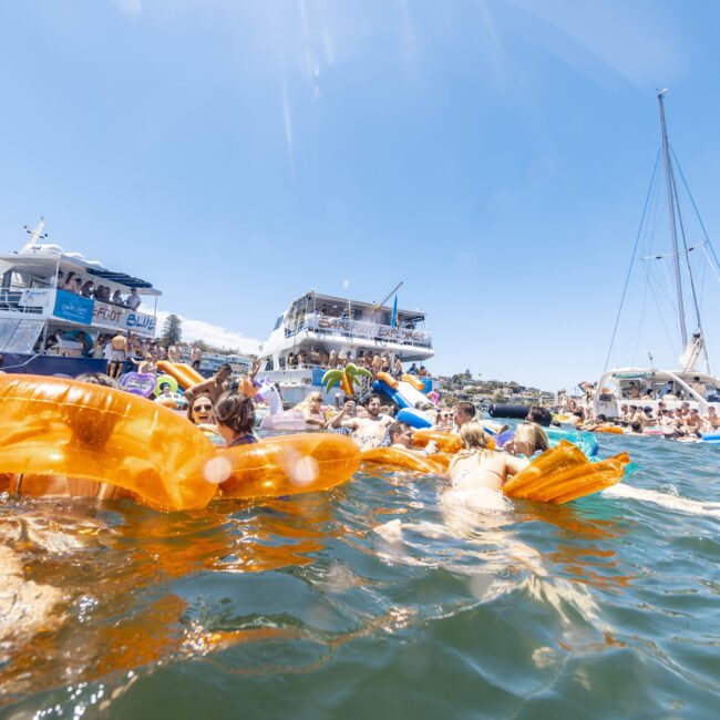 A lively scene of a boat party with numerous people enjoying the water on colorful inflatable floats. Several yachts and boats are docked nearby, with more partygoers on board. The clear, sunny sky adds to the festive atmosphere, making it a perfect day for celebration and fun on the water.