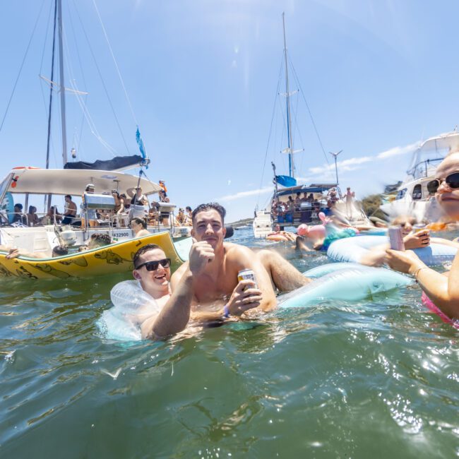 People are floating on inflatables and swimming in the water near anchored boats during a sunny day. Some are holding drinks and chatting. The background shows a boat with a visible yacht society logo, adding an air of sophistication. The atmosphere is lively and sociable.