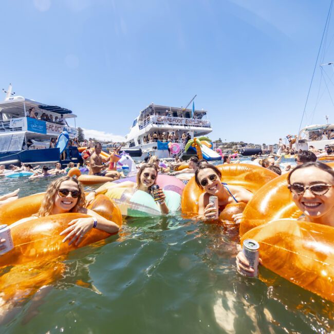 A group of people are enjoying a yacht party with several boats visible in the background. The focal point is on four women in the water, holding drinks and lounging on orange inflatable rings, smiling at the camera under a sunny blue sky, creating a picturesque summer scene.