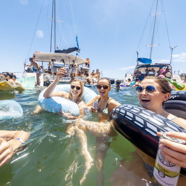 A group of people enjoy a sunny day in the water, surrounded by boats. Some float on inflatable tubes while others swim. Three women in the foreground laugh and raise drinks, showing a festive and vibrant atmosphere. The clear sky adds to the lively, joyful scene at this bustling marina.