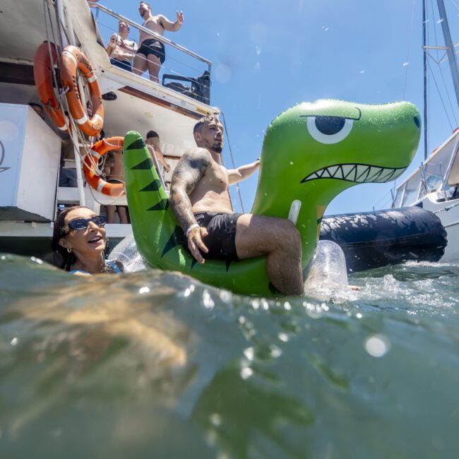 A man sits on an inflatable dinosaur float in the water while a woman swims nearby. In the background, several people on a boat cheer and wave. The sun is shining brightly, enhancing the fun, party atmosphere of this Yacht Social Club event.