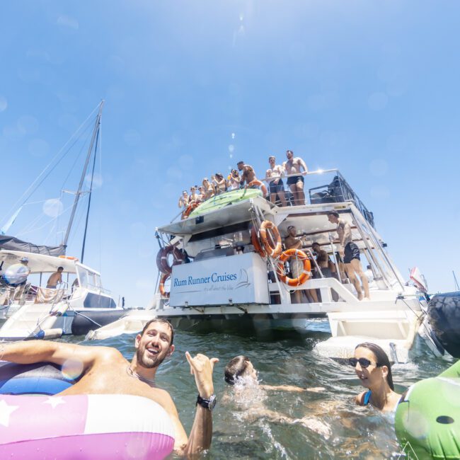 A group of people are enjoying a sunny day on the water. Some are on the deck of a boat named "Rum Runner Cruises," while others swim and float on inflatables nearby, savoring the refreshing breeze. The atmosphere is lively with clear blue skies overhead.