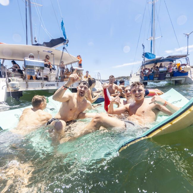 A group of people enjoying a sunny day in the water, surrounded by yachts. Some are sitting on floating mats and cheering, while others are swimming. The atmosphere is festive and joyful, with clear blue skies in the background.