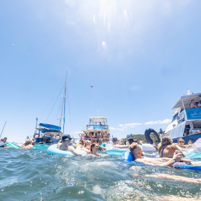 A large group of people relaxing on floating devices in the water near several boats under a clear, sunny sky. The scene is lively with people enjoying the summer day; some are sitting on yachts and others are floating close by.