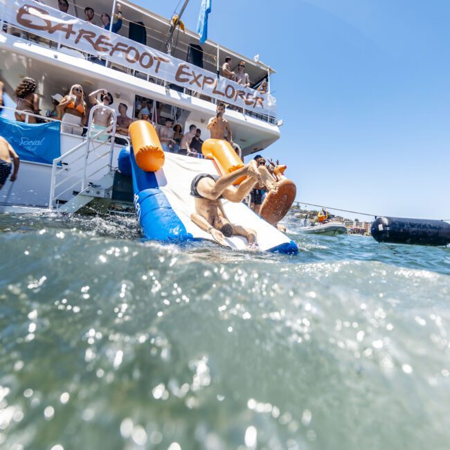 A group of people enjoy a sunny day on a boat named "Extrefot Explorer." Some are sunbathing, while others are jumping into the water or sliding down an inflatable slide attached to the boat. The water is choppy, and the sky is crystal clear, making for an exhilarating adventure.