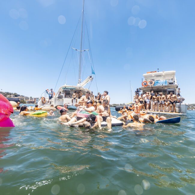 A group of friends enjoys a sunny day on the water, surrounded by boats and colorful inflatables. Some float lazily, while others relax on boats. The scene is lively under clear blue skies, with the Yacht Social Club logo visible in the corner.