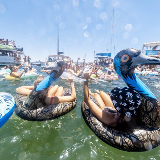 A lively scene on the water shows people enjoying a sunny day on inflatable pool floats. The focus is on two flamingo-shaped floats, each occupied by happy individuals, while boats filled with more revelers can be seen in the background under clear blue skies.