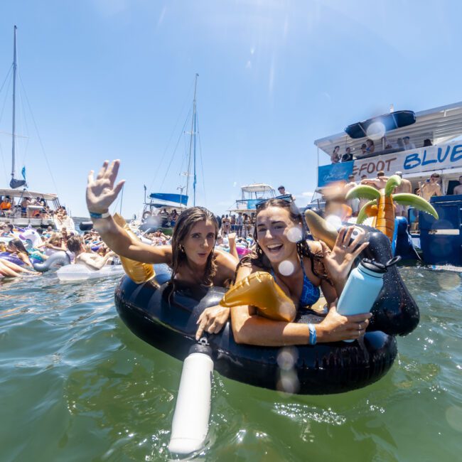 Two women float on inflatable tubes in the water, smiling and waving. They are surrounded by other people on floats and boats under a clear blue sky. A boat labeled "Foot Blue" is in the background, and a logo in the bottom right reads "Yacht Social Club." The scene is vibrant and inviting.