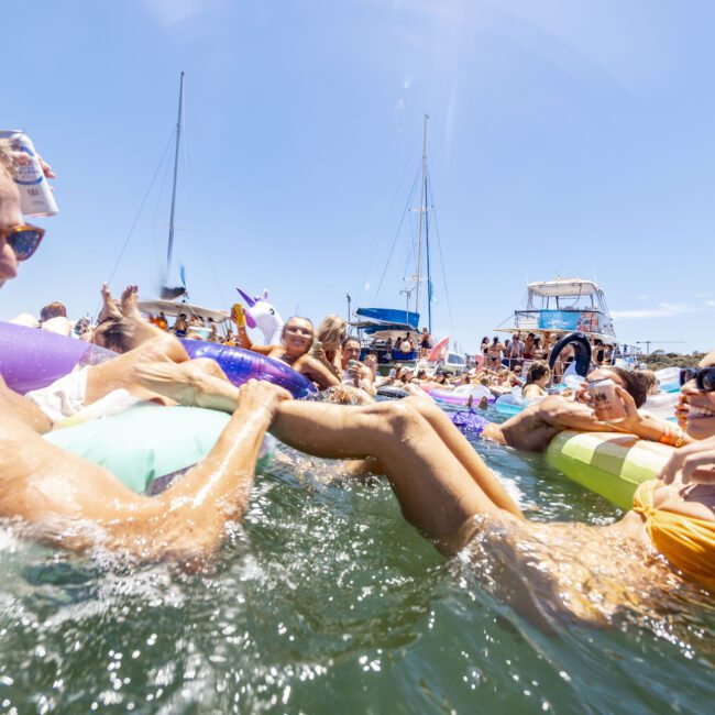 A lively group of people enjoying a sunny day on the water, floating on various inflatables near several anchored yachts. Two people in the foreground laugh and splash, while others behind them relax and socialize. The scene exudes fun, teamwork, and camaraderie.