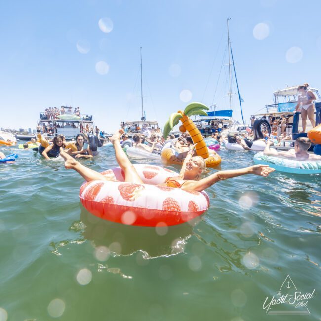 A group of people enjoy a sunny day on the water, floating on various inflatable tubes, including a woman relaxing on a strawberry-shaped float. Boats are anchored nearby, and there is a festive atmosphere with clear skies and shimmering water droplets, enhancing the overall vivid scene.