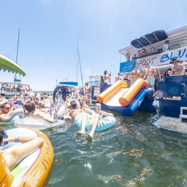 A lively scene of people enjoying a yacht party on the water. Many are floating on inflatable pool toys like a palm tree and a unicorn. Others are on the yacht, mingling excitedly and using the slides to enter the water. The day is gloriously sunny with a clear blue sky.