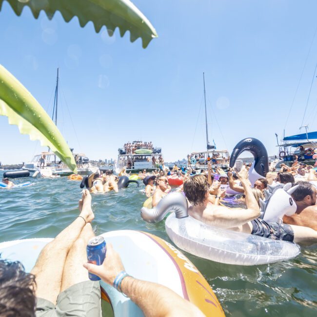 People relaxing on inflatable floats in a crowded lake, surrounded by other colorful inflatables and boats on a sunny day. A mix of paddle boards, unicorn-shaped floats, and tubes dot the water. Some people hold drinks, creating a vibrant, festive atmosphere.