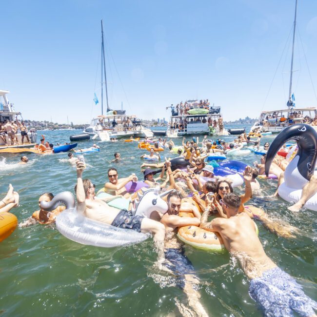 A lively group of people are enjoying a party on the water, floating on various inflatable rafts and swim rings shaped like unicorns, flamingos, and other animals. Several yachts in the background host more partygoers under a clear blue sky. A logo reads "Yacht Social Club.