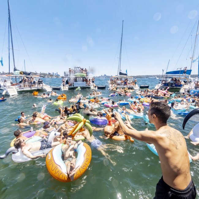 A lively scene of a yacht social gathering on a sunny day. Numerous people enjoy themselves in the water on colorful inflatables, including a unicorn, a slice of pizza, and a rainbow ring. Several boats from The Yacht Social Club Sydney Boat Hire are anchored nearby, providing music and a festive atmosphere.