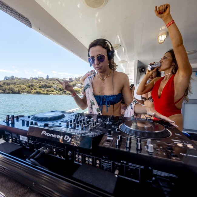A woman wearing sunglasses and a bikini operates DJ equipment on a boat under a clear sky. Another woman in a red bikini dances energetically behind her. They are on a waterfront, surrounded by a scenic view of water and lush greenery, creating an oceanic ambiance.