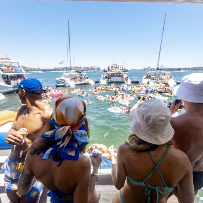 A group of people on a boat look out at numerous boats and people floating in the water on inflatables. Some wear swimsuits, hats, and sunglasses, while holding drinks. The lively scene depicts a sunlit aquatic gathering with clear skies in the background during a perfect summer day.
