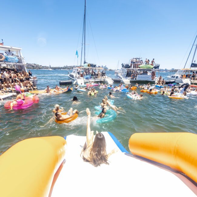 A lively scene of a boat party on a sunny day features many people floating and swimming in the water. Inflatable boats and colorful floats are scattered around. Multiple yachts are anchored nearby as drones capture the fun. People are enjoying the summer vibes and socializing.