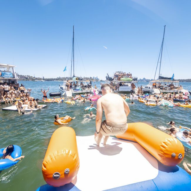 A man prepares to glide down an inflatable water slide into a lake, surrounded by people on vibrant floaties and several anchored yachts. The clear sky and daytime setting create a lively atmosphere, with everyone enjoying the water on this sunny day.