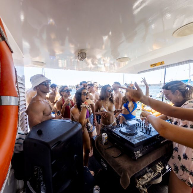 A group of people in swimwear are dancing and enjoying music on a boat. A DJ is playing tunes on a mixer at the center of the scene. Various people are holding drinks and wearing sunglasses and hats. The Yacht Social Club Event Boat Charters ensures everyone has a great time.