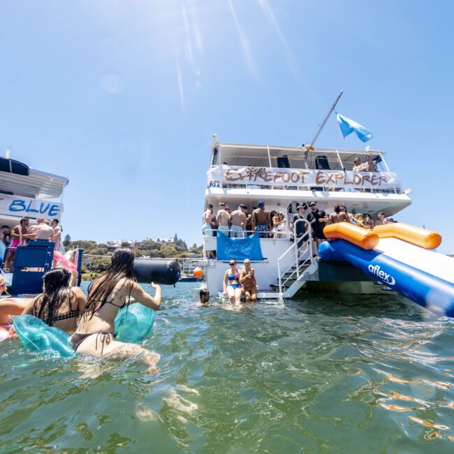 People are enjoying a sunny day on the water with two large boats. One boat features a slide into the water while the other hosts a group on top. Several individuals are swimming near the boats, and some are lounging on vibrant floats. The atmosphere is lively and festive, adding to the summertime fun.