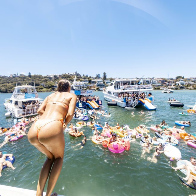 A person in a bikini is preparing to jump off a boat into the water. The water is filled with multiple people on colorful floatation devices and paddleboards. Several boats are anchored nearby, and the background shows a shoreline with houses and greenery.