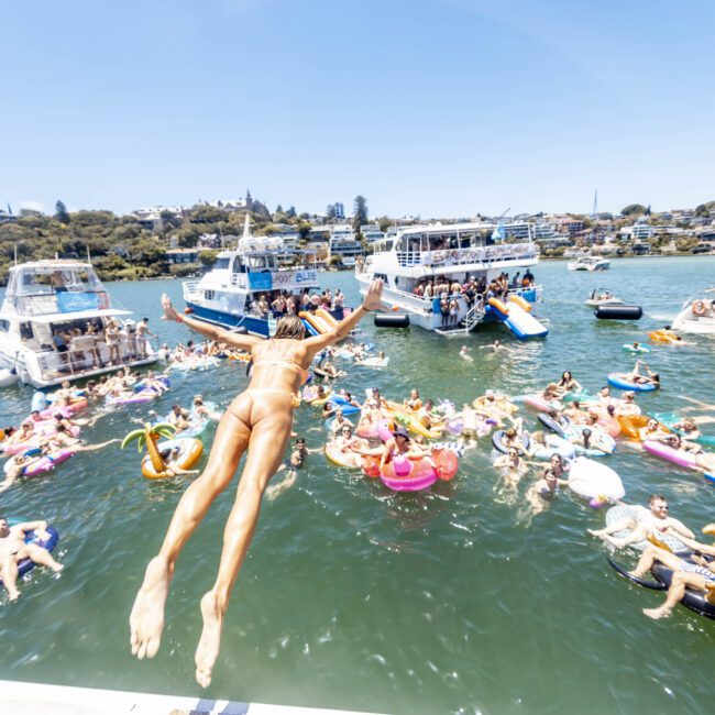 A person in a bikini dives into a lively lake from a boat. Many people are in the water on floats and inflatables, enjoying the sunny day. Several boats are anchored nearby, with people relaxing on them. The background includes trees and some picturesque houses.