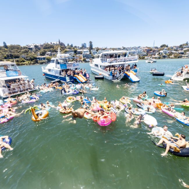 A lively scene of people enjoying a sunny day on the water, with some lounging on inflatable pools, floats, and boats. Multiple yachts are docked nearby, and numerous individuals are swimming and relaxing in the clear water.