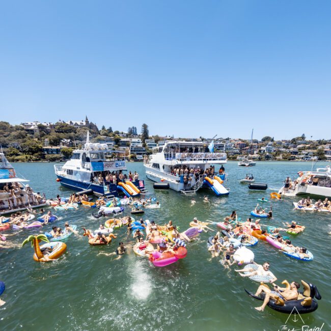 A lively scene of people enjoying a sunny day on the water, floating on colorful inflatables near several anchored boats. The shoreline villas are seen in the background. Some individuals are swimming while others relax. A logo says "Yacht Social Club" in the bottom right corner.