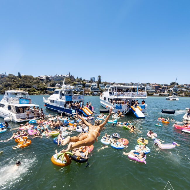A lively crowd enjoys a sunny day on the water, with several people on colorful floaties and a boat party in the background. A person is mid-air, diving into the lake, adding to the festive atmosphere. Boats and waterfront houses complete this picturesque scene.