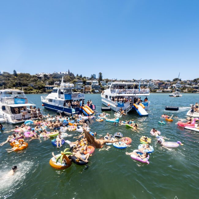 A lively scene of people enjoying a sunny day on a lake, surrounded by several boats. Many individuals float on colorful inner tubes and inflatables. The background showcases lush greenery and distant houses. The Yacht Social Club logo is visible in the corner, highlighting the joyous atmosphere.
