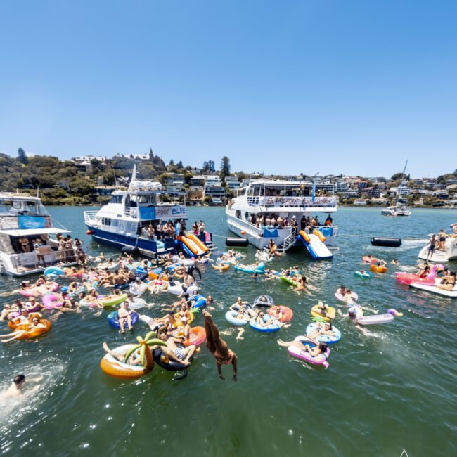 A lively water party with numerous people on inflatable floats in a lake. Several boats are anchored nearby, with attendees swimming and enjoying the sunny weather. The image features "The Yacht Social Club" logo in the bottom right corner, capturing a vibrant and social atmosphere.