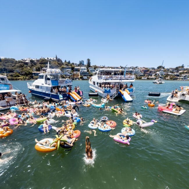 People on boats and colorful inflatables gather at a lively yacht social event on a sunny day. The scene, set in a bay with green hills and houses in the background, is vibrant. Swimmers in the water enjoy the festivities under clear blue skies.
