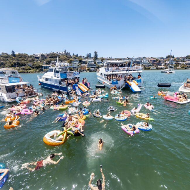 A vibrant scene of people enjoying a sunny day on the water, surrounded by boats. Many float on colorful inflatables and rafts, swimming and socializing under the clear blue sky. In the background, houses and trees line the shore. Logo in the bottom right corner reads "Yacht Social Club".